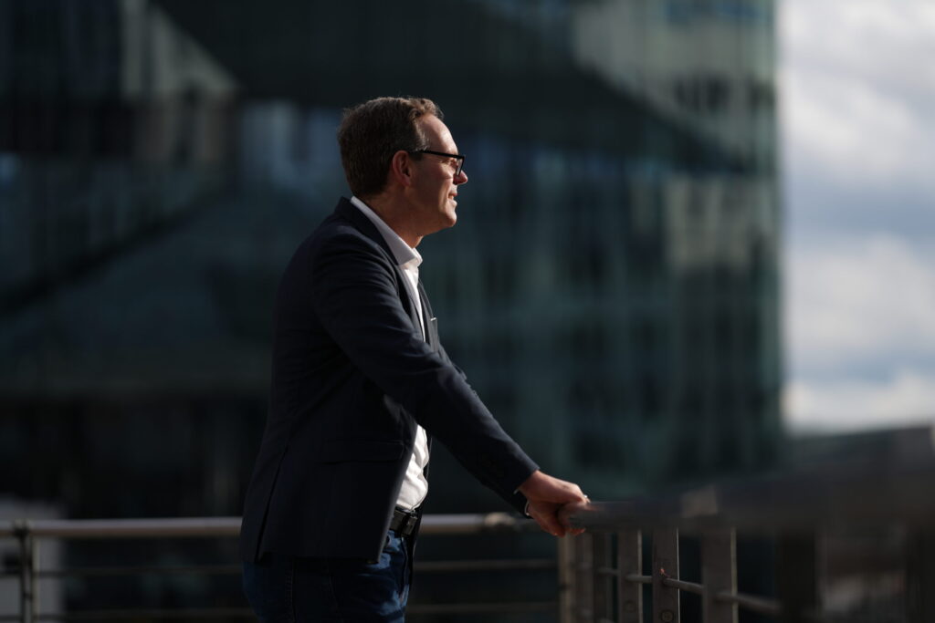 Man in a suit standing outdoors, leaning on a railing, looking into the distance with a cityscape background