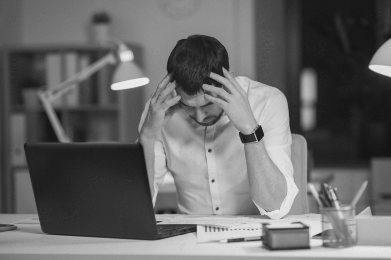 Stressed man holding head at desk with laptop and documents
